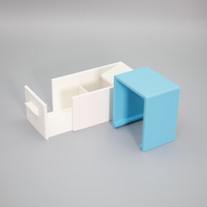 K&ouml;nnte beinhalten: Zwei minimalistische Aufbewahrungsbeh&auml;lter. Einer ist wei&szlig; mit geteiltem Innenraum und einer Schublade. Der andere ist eine kleine, hellblaue, offene Box. Beide bestehen aus einem matten Material.