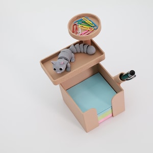 Può includere: Un organizer da scrivania beige con un coperchio a forma di gatto, un piccolo vassoio con graffette colorate e un portapenne. L'organizer contiene una pila di post-it pastello. Il gatto è grigio con grandi occhi.