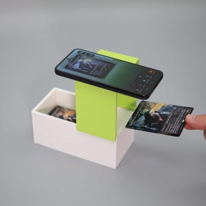 K&ouml;nnte beinhalten: Ein Smartphone, das eine Kartenspieloberfl&auml;che anzeigt, ruht auf einem hellgr&uuml;nen St&auml;nder &uuml;ber einer wei&szlig;en Box. Eine Karte wird in der Hand gehalten, und andere Karten sind in der Box sichtbar. Der Telefonbildschirm zeigt ein Kartenspiel.