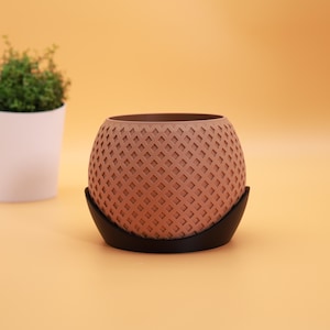 Op de afbeelding: Een bruine, bolvormige bloempot met een rasterpatroon, rustend op een zwarte, gebogen basis. De pot is leeg en de achtergrond is effen lichtoranje. Een kleine witte pot met een groene plant is zichtbaar op de achtergrond.