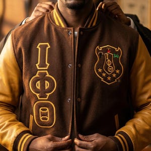 May include: Brown and gold varsity jacket with leather sleeves. The jacket features the Greek letters I, Φ, and O in gold, along with a crest emblem. The jacket has a button-up front and striped cuffs.