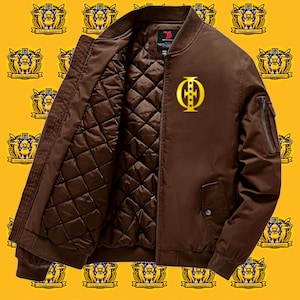 Könnte beinhalten: Braune Bomberjacke mit gestepptem Innenfutter und goldenem Emblem auf der Brust. Die Jacke hat einen Reißverschluss, einen gerippten Kragen und eine Tasche am linken Ärmel. Der Hintergrund ist gelb mit einem sich wiederholenden Wappenmuster.