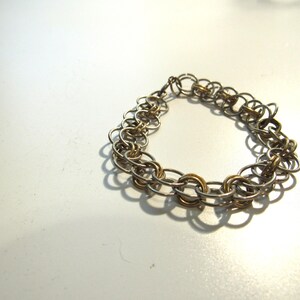 May include: A silver and gold-toned chain bracelet. The bracelet is composed of multiple interlocking oval links. Some links are silver, while others are gold. The bracelet is resting on a white surface.