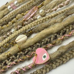 May include: Close-up of handmade dreadlocks in various shades of brown and blonde. Adornments include pearls, pink crystals, a seashell, and a pink heart charm with an eye design. Braided and wrapped sections add texture.