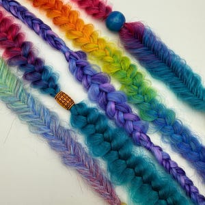 May include: Close-up of several colorful braided hair extensions in a rainbow of hues. Each braid features a different color scheme, including pink, orange, yellow, green, blue, and purple. Some braids have decorative beads at the ends.