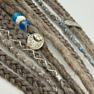 May include: Close-up of brown and gray dreadlocks with blue and white beads, silver charms, and braided accents. The dreadlocks are styled with silver thread and a silver wing charm. The dreadlocks are a natural color.