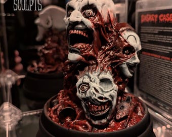 May include: A sculpted figurine featuring three pale, grotesque faces with blood-red textures and exposed teeth, set atop a black circular base. The text NEFARIOUS SCULPTS is visible in the upper left corner, and a small rat figure is integrated into the base.