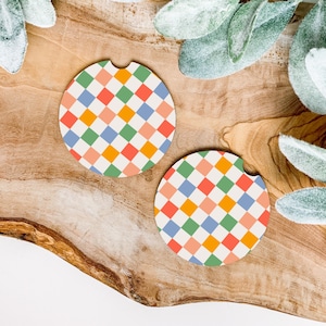 Puede incluir: Dos posavasos redondos para coche con un colorido diseño a cuadros en naranja, rosa, verde y azul. Se colocan sobre una superficie de madera con acentos de hojas verdes, creando un accesorio vibrante y decorativo.