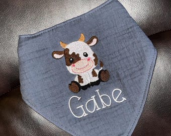 May include: A blue bandana-style baby bib with an embroidered cartoon cow. The cow is white, brown, and black, with small yellow horns. The name "Gabe" is embroidered in white script below the cow.