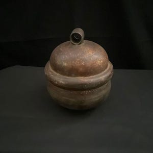 Könnte beinhalten: Ein antiker Metallbehälter mit Deckel und kleinem Schlaufengriff. Der Behälter hat eine abgerundete Form mit dunkler, gealterter Bronzeoberfläche, was darauf hindeutet, dass er schon lange existiert. Das Design ist einfach und funktional.