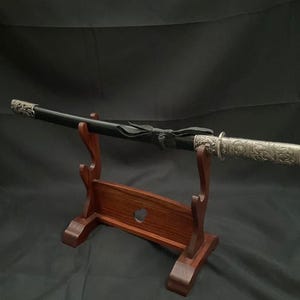 以下が含まれることがあります： 黒い鞘と装飾的な銀色の柄が付いた日本の刀、刀が木製のディスプレイスタンドに置かれています。スタンドは濃い茶色で、ハート型の切り抜きがあります。刀は伝統的な武器です。