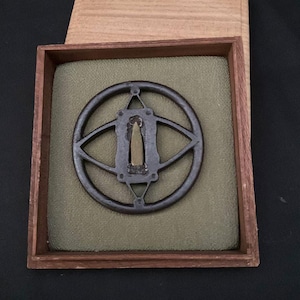 May include: A dark iron tsuba featuring a circular outer frame with an internal geometric design and a central brass inlay, presented inside a square wooden box with a light green fabric lining and a separate wooden lid resting behind the base.