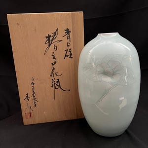 以下が含まれることがあります： 淡い緑色のセラミック花瓶と木箱。花瓶は滑らかな表面と繊細な花のイラストが特徴です。木箱には日本の書道があります。