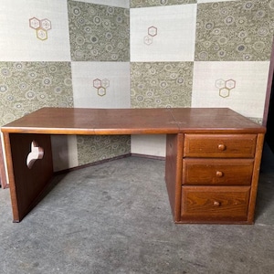 Könnte beinhalten: Ein Holzschreibtisch mit warmbraunem Finish. Der Schreibtisch hat eine rechteckige Tischplatte, eine Seite mit dekorativem Ausschnitt und drei Schubladen. Der Hintergrund ist eine gemusterte Wand.