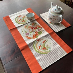 以下が含まれることがあります： 白とオレンジのストライプ柄のテーブルランナーに、刺繍された花柄が施されています。ランナーは濃い木製のテーブルの上に置かれ、白いティーカップと龍の模様のティーポットが置かれています。