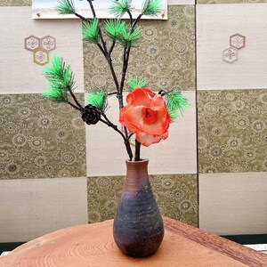 以下が含まれることがあります： 小さな、濃い青と茶色の花瓶には、緑の針、黒い松ぼっくり、鮮やかなオレンジ色の花が付いた枝が入っています。 花瓶は、模様のあるパネルを背景に、自然な木目模様の木製のテーブルの上に置かれています。