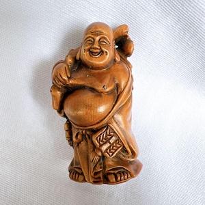 May include: A wooden figurine of a smiling, round-bellied Buddha holding a bag of money in one hand and a staff in the other. The figurine is carved in a traditional style and has a warm brown color.