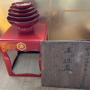 May include: A stack of red lacquered serving bowls with gold trim, atop a matching red stand with a gold emblem. A wooden box with Japanese text sits to the side. The bowls are of varying sizes.