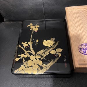 May include: Black lacquered box with a gold painted bird and floral design. The box has a glossy finish and a rectangular shape. A wooden box is partially visible on the right side. The bird is perched on a branch with gold leaves and red berries.