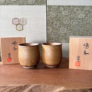 以下が含まれることがあります： 温かみのあるアースブラウンの釉薬を施した、手作りの質感のあるセラミックティーカップ2個。カップは、それぞれ日本語の文字が入った2つの木箱と一緒に展示されています。背景には模様のある壁があります。