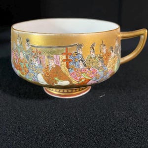 May include: A gold teacup with a detailed hand-painted scene of people in traditional Japanese clothing. The cup has a white interior and a gold handle.