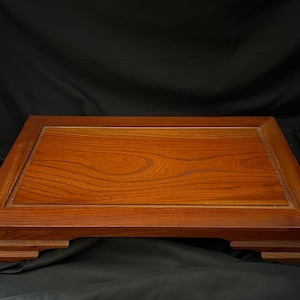May include: A rectangular wooden display stand featuring a warm, reddish-brown wood grain finish. The platform has a raised border and is supported by four stepped, tiered feet at each corner, creating a low-profile, elevated base structure.