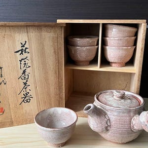 以下が含まれることがあります： サイドハンドル付きの急須と5つの湯呑みがセットになった陶器の茶器セット。表面は質感のある淡いピンクベージュの釉薬で仕上げられています。木製の収納箱には、黒い日本語の書道と赤い印章が施されています。