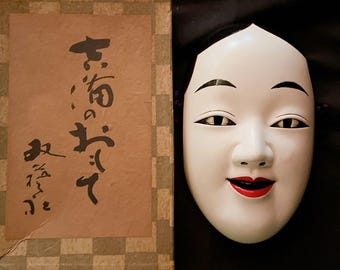 Könnte beinhalten: Eine weiße Noh-Maske mit einem lächelnden Ausdruck, roten Lippen und schwarzen Augen. Die Maske befindet sich in einer Holzkiste mit japanischen Schriftzeichen auf der Vorderseite.