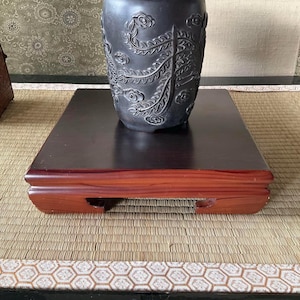 以下が含まれることがあります： 複雑な花と鳥のデザインが施された黒い陶器の花瓶が、赤い木製の縁取りのある暗い木製の台の上に置かれています。台は長方形の天板とわずかに湾曲したベースを持っています。