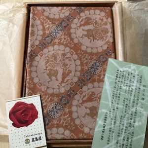 以下が含まれることがあります： 茶色とベージュの色合いの模様入りの布カバーが付いた装飾箱。円形のデザインには神話上の生き物が描かれています。赤いバラと日本語のテキストが入った小さなカードが見えます。