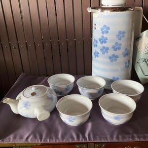 以下が含まれることがあります： 青い花柄の白いティーポットと白いティーカップ6個。ティーポットは注ぎ口と取っ手があります。ティーカップはすべて同じサイズで丸みを帯びています。ティーカップとティーポットは紫色のテーブルクロスの上に置かれています。背景には青い花柄の白い魔法瓶があります。