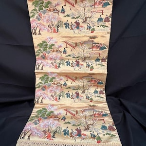 May include: A rectangular, golden-hued textile with a repeating Japanese landscape pattern. The design features figures, bridges, and buildings, with cherry blossom trees. The top and bottom edges have a beige fringe.