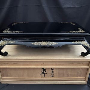 May include: A black lacquered wooden display stand with gold floral accents. The stand has a rectangular top and curved legs. It sits atop a light-colored wooden box with Japanese characters.