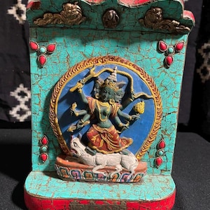 以下が含まれることがあります： ターコイズと赤のウッド製の祠。カラフルな神様の像が複数の手と白い動物を従えて立っています。神様は青と金の輪郭で囲まれています。祠はターコイズと赤の石で飾られています。