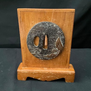 Peut inclure: Un support de présentation rectangulaire en bois avec une tsuba métallique circulaire centrée. La tsuba présente une finition sombre et texturée avec un motif d'aile métallique et deux découpes. La base en bois clair a un grain naturel.