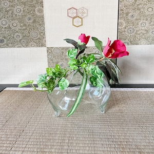 以下が含まれることがあります： 3本の脚が付いた透明なガラスの花瓶に、緑の葉の植物とピンクの椿の花が2輪入っています