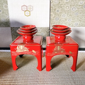 May include: Two red lacquered stands with gold painted designs, each topped with a stack of four red and gold bowls. The stands have four legs and are decorated with a scene of a boat on water.
