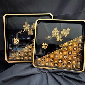 以下が含まれることがあります： 金色の縁取りと装飾的な金箔のデザインが施された、2つの黒い漆塗りのトレイ。各トレイは、小さな正方形のパターンと金箔の枝のデザインが施された対角線セクションを備えています。トレイは正方形です。