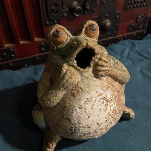 以下が含まれることがあります： 口を大きく開けた緑と茶色の釉薬が施された白いアクセントのあるセラミックの蛙の置物。蛙は茶色の瞳を持つ大きく丸い目を持っている。