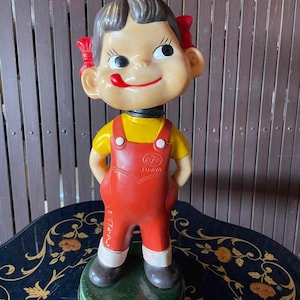Könnte beinhalten: Eine vintage, rot-gelbe Plastikfigur eines Mädchens mit einer roten Schleife im Haar. Die Figur hält ihre Hände in die Hüften und hat ein lächelndes Gesicht. Die Figur steht auf einem grünen Sockel mit dem Text "フジヤ" in japanischen Schriftzeichen.