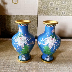 以下が含まれることがあります： 金色の縁と底を持つ2つの青い七宝焼きの花瓶。各花瓶は、白とピンクの花、緑の葉、複雑なディテールを備えた花の模様が特徴です。花瓶は織りのマットの上に置かれています。