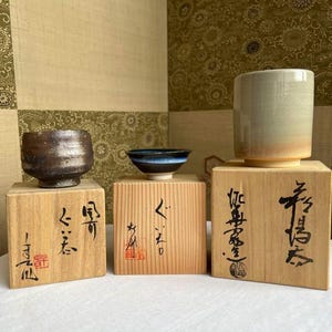 以下が含まれることがあります： 木製の台座に置かれた、3つの陶磁器製の茶碗。茶碗は、茶色、黒、白など、さまざまなスタイルと色で構成されています。台座はすべて明るい木で作られており、日本語が書かれています。