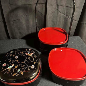 May include: A three-tiered black lacquered bento box set with red interior and a black metal carrying handle. The top tier features a floral design with birds in gold and black.