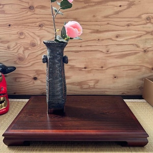 以下が含まれることがあります： 2つのハンドルが付いた濃い茶色の織りの花瓶には、ピンクの花と緑の葉が付いた花の枝が入っています。 花瓶は、縁が盛り上がった濃い木製の正方形のトレイの上に置かれています。 背景は明るい木製のパネルです。