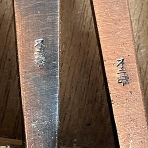 Może przedstawiać: Dwa metalowe uchwyty sztućc&oacute;w w odcieniu miedzi ustawione pionowo. Każdy uchwyt ma szczotkowaną, teksturowaną powierzchnię i jest wytłoczony identycznymi japońskimi znakami kanji w zagłębionej, ciemnej czcionce na środku trzonka.