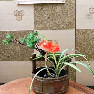 以下が含まれることがあります： 小さな木製のバケツ型の容器に、鮮やかなオレンジ色の花と緑の葉をあしらった日本の生け花。木製の表面に配置され、模様のある背景と書道が添えられています。