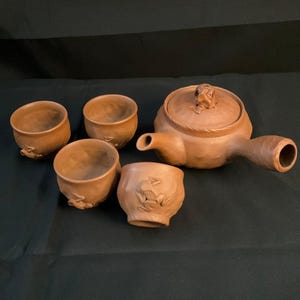 May include: A brown ceramic tea set, comprising a teapot with a lid and four cups. The teapot has a long handle and spout. The cups and teapot have a textured surface and a rustic appearance. The cups feature a frog design.