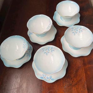 以下が含まれることがあります： 5つの磁器製のティーカップとソーサーのセット。カップとソーサーは白で、水色のトリムと金の縁取りが施されています。ソーサーはスカラップエッジで、繊細な青い葉のデザインが施されています。