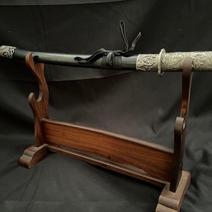 以下が含まれることがあります： 装飾的な銀色のディテールが施された黒い剣が、木製のスタンドに置かれています。 剣の柄は黒い素材で包まれており、スタンドは濃い色の木材で作られています。 剣の刃は見えません。