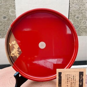 以下が含まれることがあります： 中央に白い花のエンブレムが付いた、丸い赤い漆塗りのトレイ。トレイの一部には金箔のアクセントがあります。トレイは黒いスタンドに展示されており、近くに木のカードがあります。カードには日本語のテキストが書かれています。